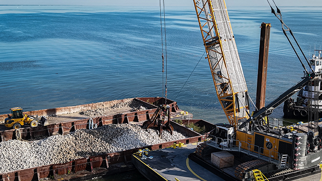 A grapple suspended from a crane lifts cobble from a barge. The crane and barge are surrounded by calm water. In the background is an empty barge with an idle front-end loader.